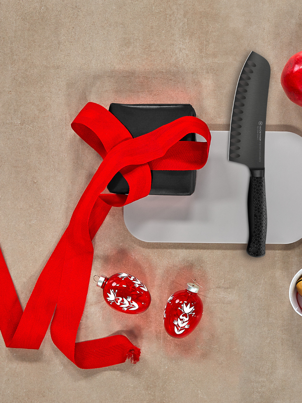 Red ribbon tied around a black object on a beige surface with a knife and red decorative items.