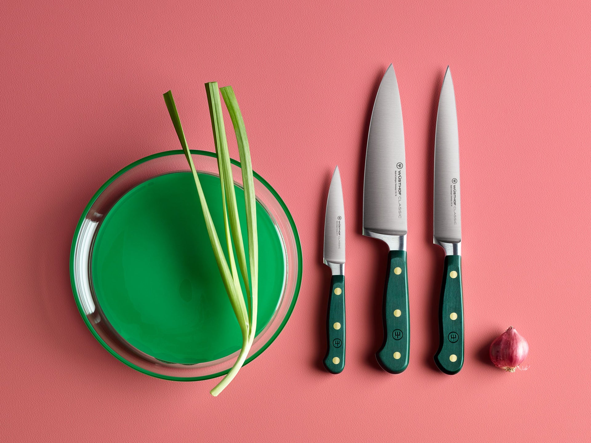 A glass bowl with green liquid and green onions, three knives with green handles, and a small red onion on a pink surface.