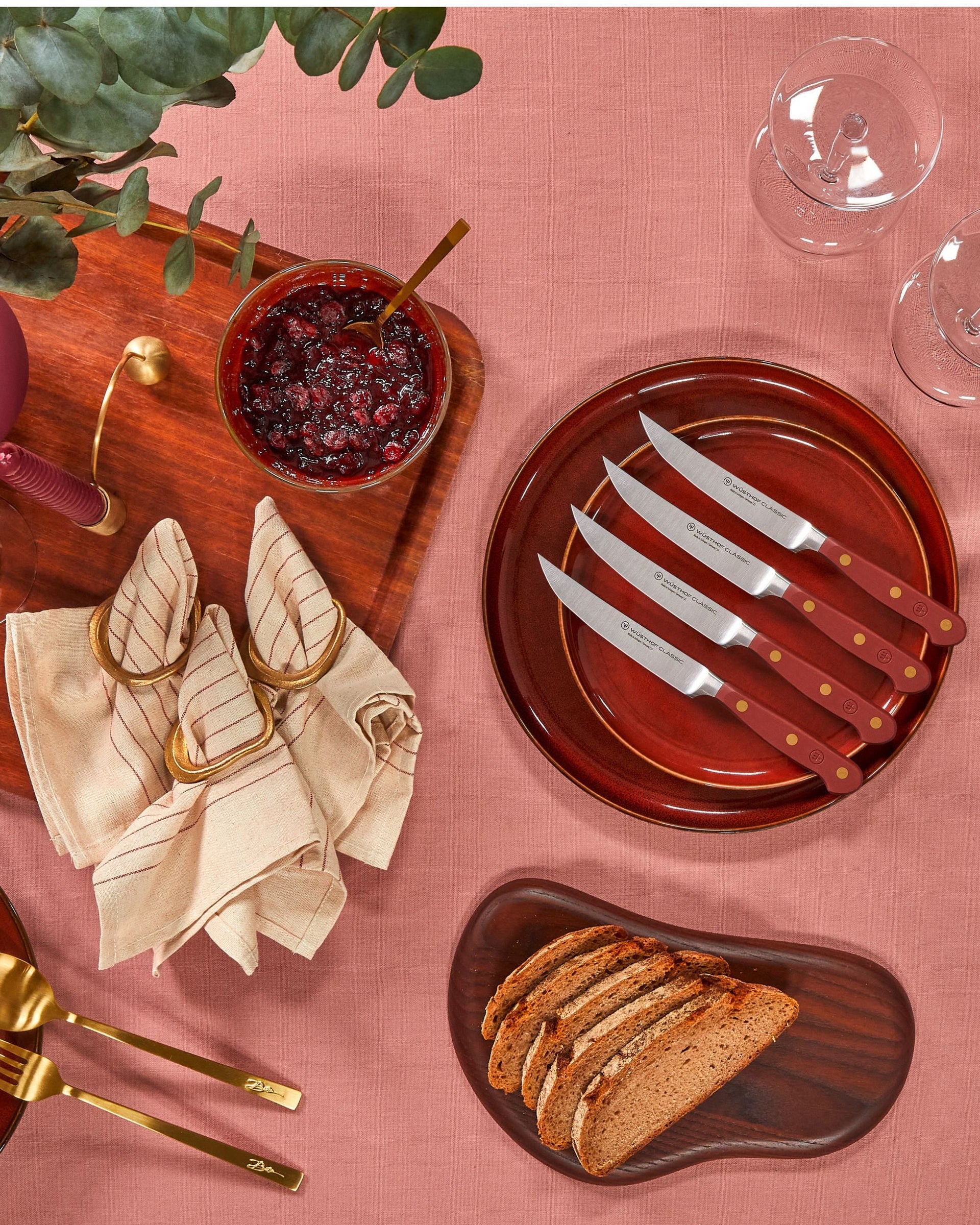 Dinner setting with red plates, knives, bread, and glasses on a pink tablecloth.