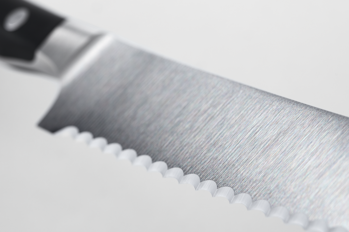 Close-up view of a serrated stainless steel knife blade with a brushed finish and part of a black handle visible against a white background.