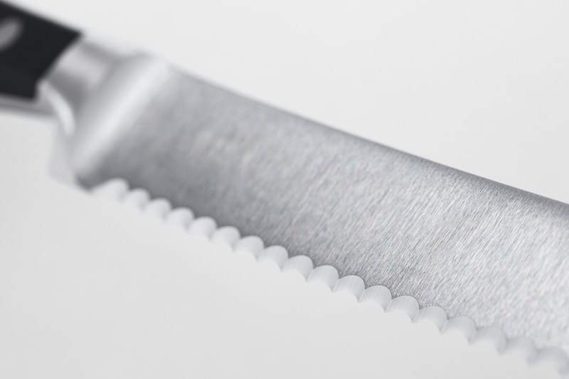 Close-up of a serrated stainless steel blade with a brushed finish and a partially visible black handle, highlighting the sharp scalloped edge.